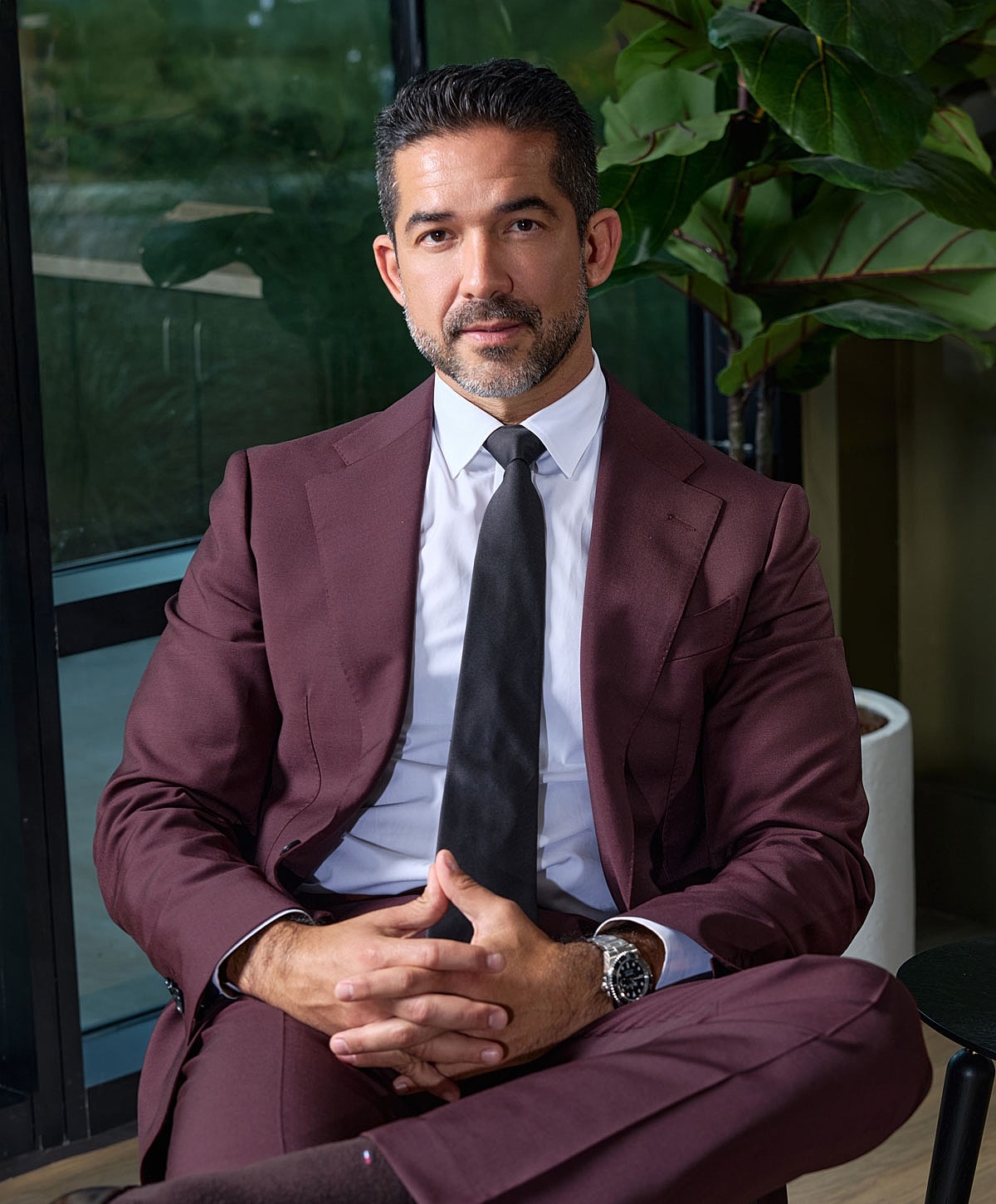 Man in burgundy suit sitting confidently indoors.