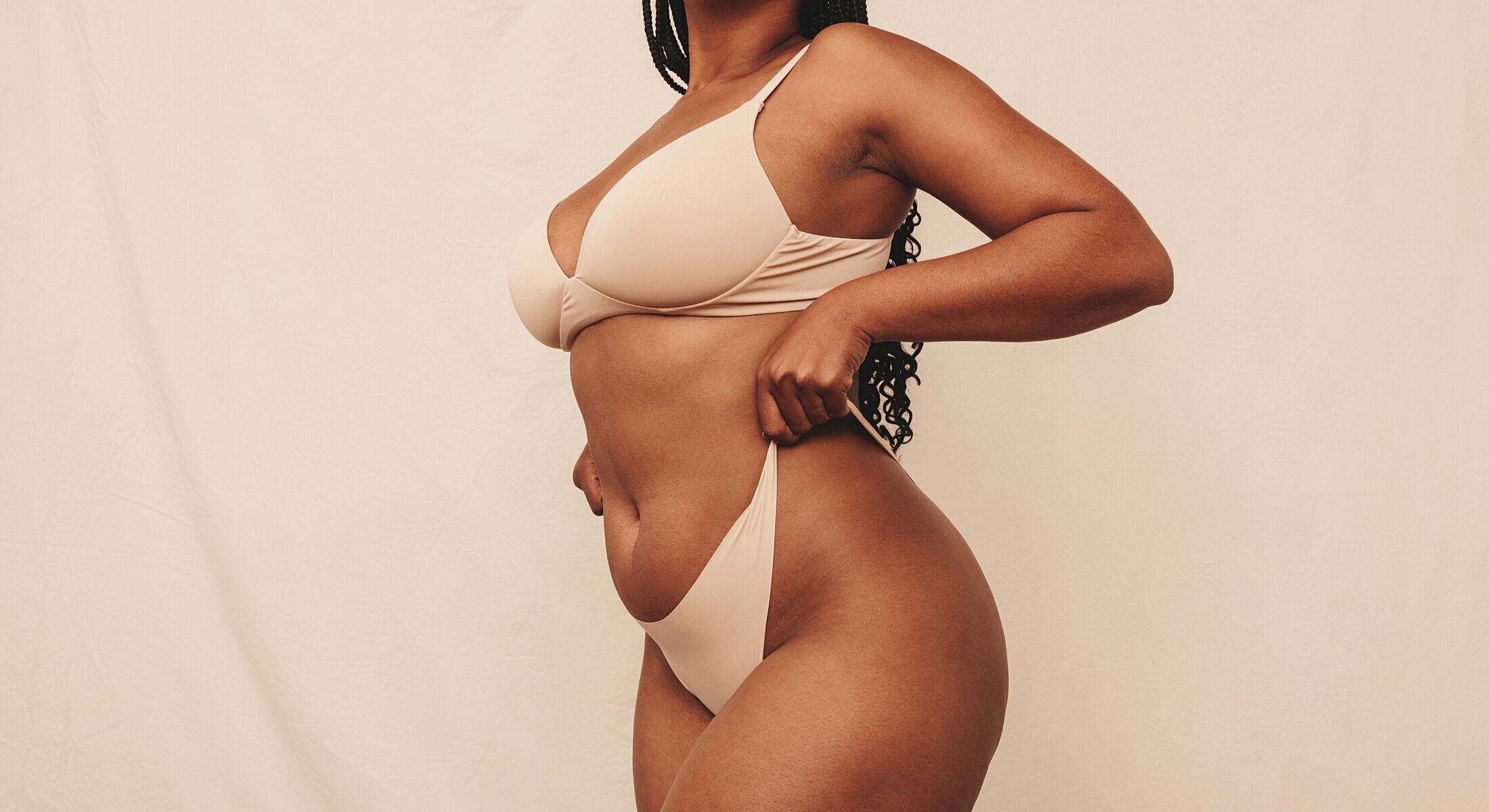 Model posing in nude lingerie against beige background.